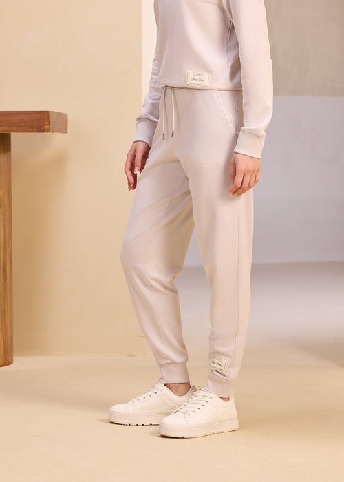 Full-length image of a person dressed in a monochrome grey lounge set featuring a drawstring hoodie and matching joggers, accessorized with white chunky sneakers, standing in a soft-hued minimalist setting.