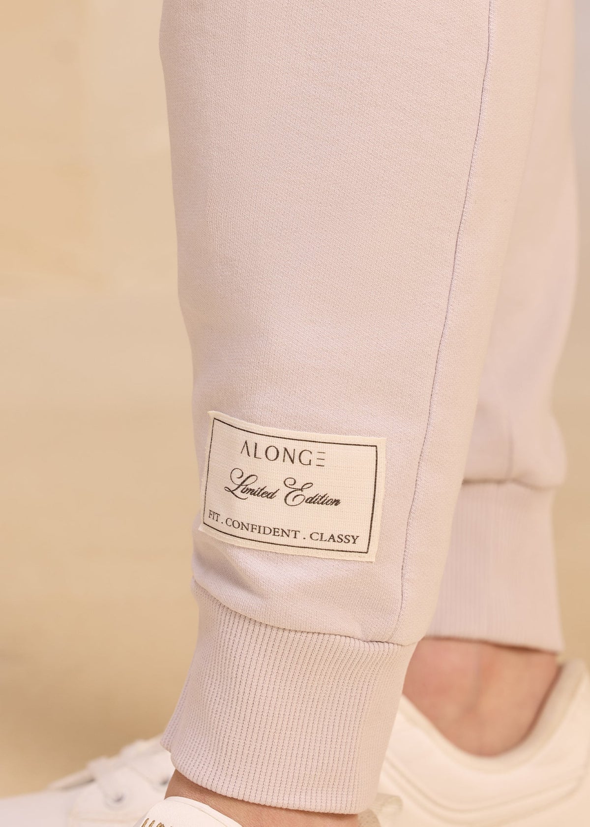 Close-up image of a grey jogger pant leg featuring a patch with the text 'ALONGE Limited Edition' and the words 'Fit. Confident. Classy,' showcasing the detailed branding in a minimalist setting.